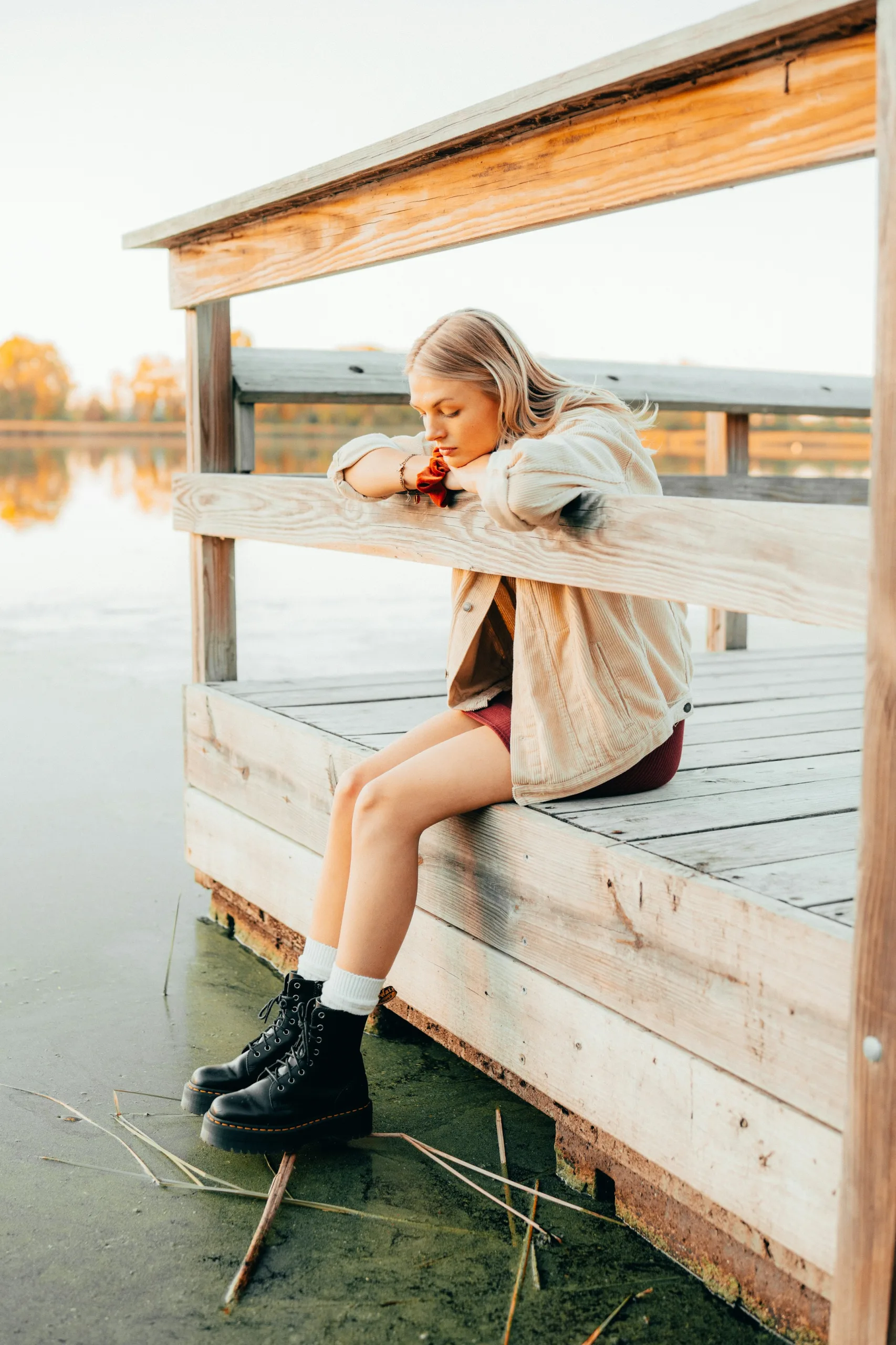 Chica sentada en un muelle de madera junto al agua, con botas negras